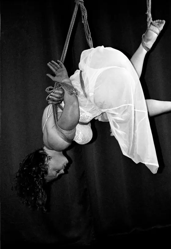 Woman suspended upside down in sheer dress and lingerie, bound with rope against a dark backdrop.
