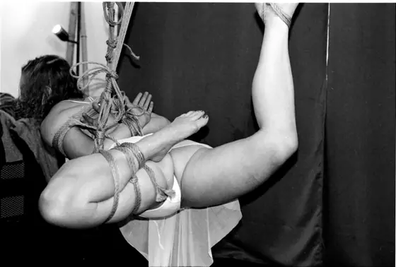 Woman suspended horizontally by intricate rope bondage against a dark backdrop, wearing white underwear and with arms and legs tightly