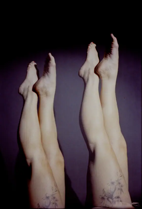 Four bare legs raised vertically against a dark background, two with visible floral tattoos on the calves.