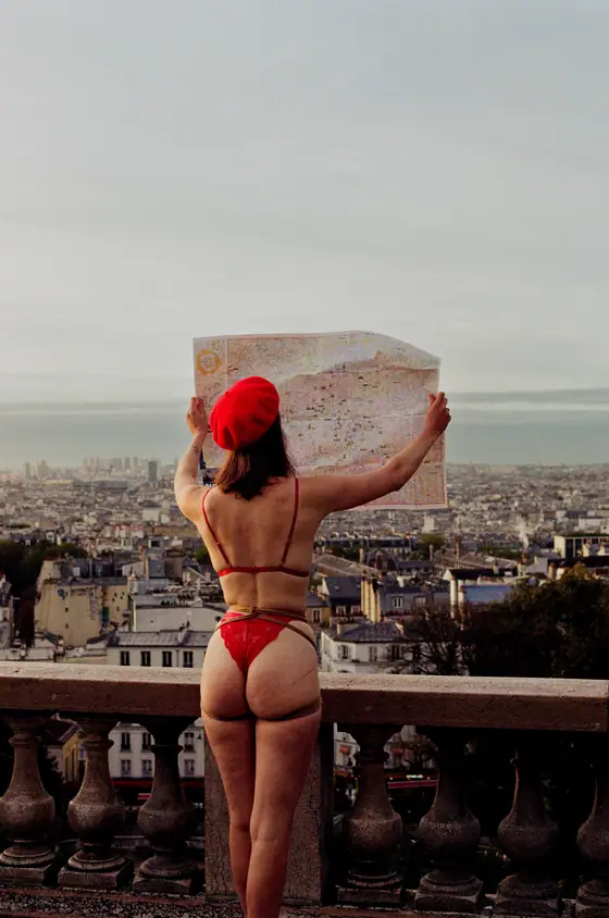 Woman in red lingerie and beret holding a map overlooking a cityscape from a stone balcony.