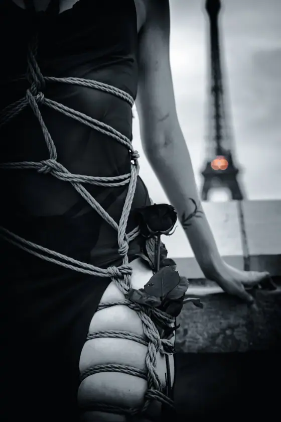Person wearing black dress tied with ropes around torso and legs, holding a black rose, with blurred Eiffel Tower in background.