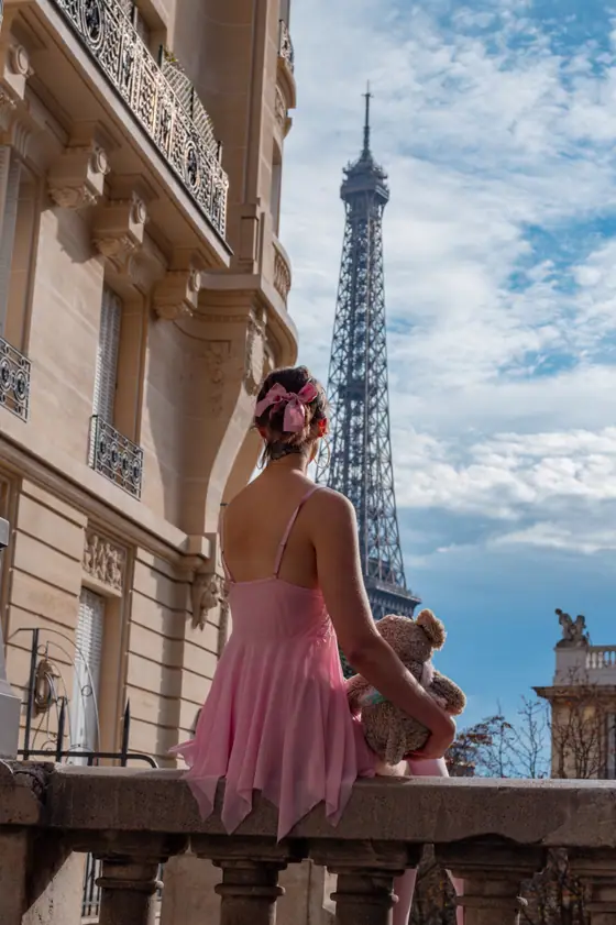 Young woman in a pink dress and bow sitting on a stone balustrade holding a teddy bear, facing the Eiffel Tower under a partly cloudy sky.
