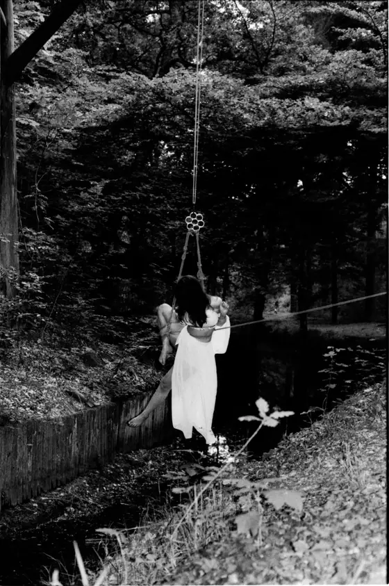 Woman in a flowing white dress suspended on a rope swing over a narrow water canal in a dense forest.