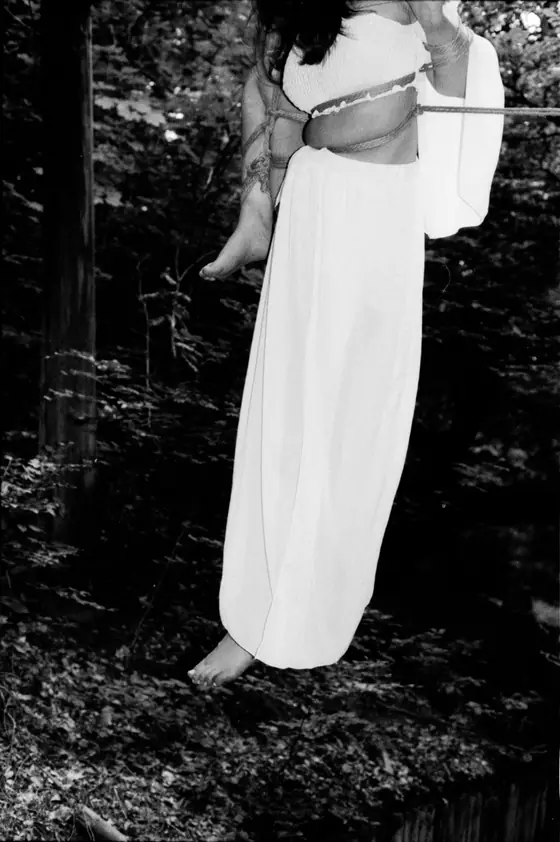 Person in long white dress suspended by ropes in forested area, black and white photo.