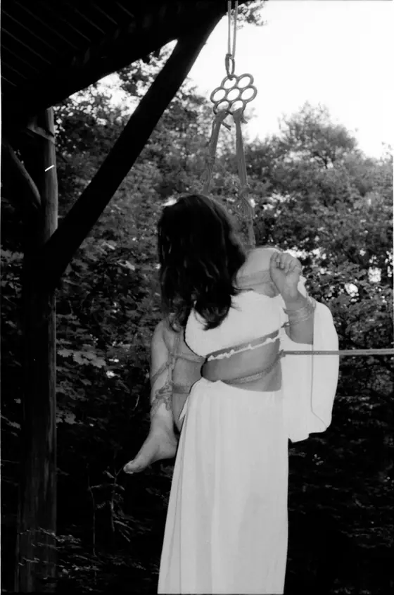 Person dressed in white with long hair suspended and bound by ropes hanging from a wooden structure in a wooded area.