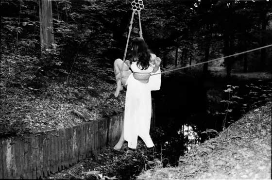 Person suspended in white clothing from ropes, hanging by a rope pulley system over a wooded area near a small body of water.