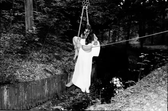 Woman in long, flowing dress suspended by rope harness above a small stream in a wooded area.
