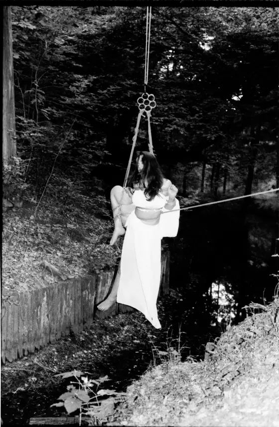 Woman in a white dress suspended by ropes above a narrow creek in a wooded area.